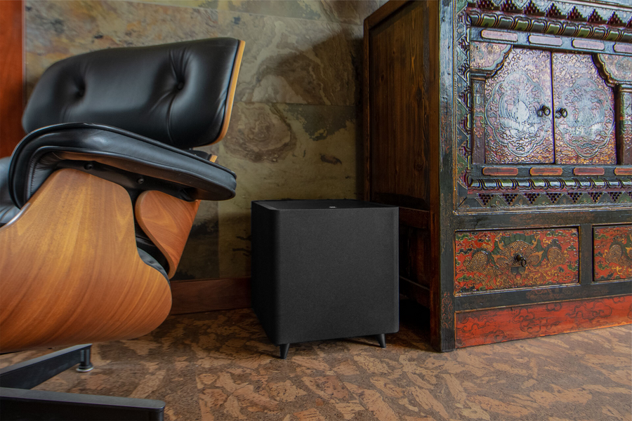 An Origin Acoustics speakers on the floor next to a midcentury-style chair and an antique dresser. 