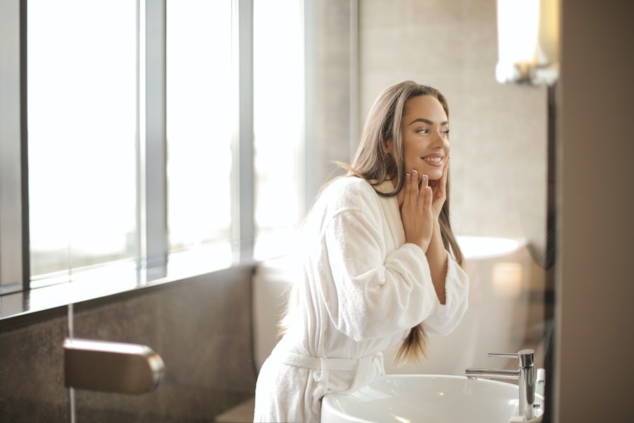 A woman smiles while looking at her skin in a luxurious bathroom.